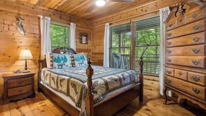 Een slaapkamer in een houten hutje, met een bed, een kaptafel, nachtkastje en een plafondventilator.