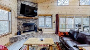 Interior d'una cabana de fusta amb una llar de pedra, un televisor fixat a la paret i un sofà de cuir.