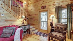 A wooden cabin interior with a staircase, a red couch, a patterned armchair, and a door leading outside. - Jack Bear's Cabin Stay (Blue Ridge)