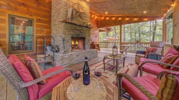 A cozy cabin interior with a stone fireplace, wicker furniture, and a large window overlooking a forest. - Jack Bear's Cabin Stay (Blue Ridge)