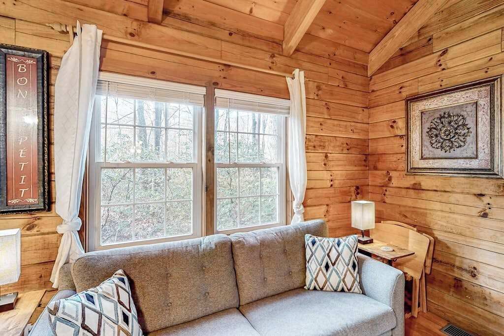 Interior de una cabaña de troncos con un sofá, dos ventanas con cortinas, un cuadro enmarcado y una mesa con una lámpara.