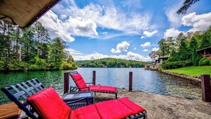 Terraza junto al lago con sillones y una mesa, con vistas a un lago tranquilo y zona boscosa.