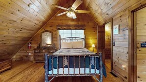 Quarto de cabana de tronco com cama, ventilador de teto, cómoda e janela com persianas.