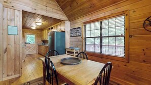 Interior de uma cabana de madeira com uma mesa de jantar, cadeiras, um frigorífico e uma janela com persianas.