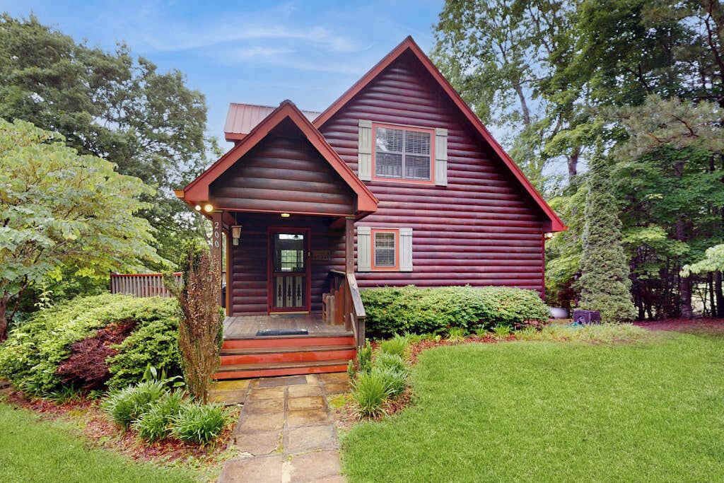A log cabin with a front porch, surrounded by lush greenery and trees.