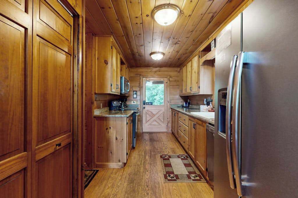 Interior de una cabaña de madera con cocina, armarios de madera, un refrigerador y una puerta que da al exterior.