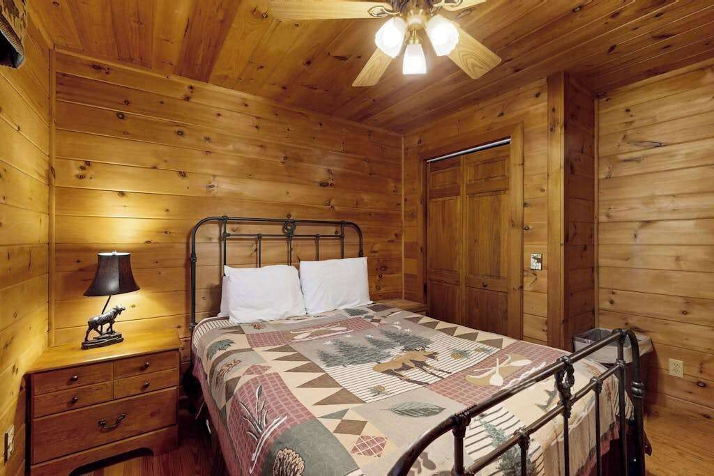 A wooden cabin bedroom with a bed, nightstand, lamp, and a ceiling fan.