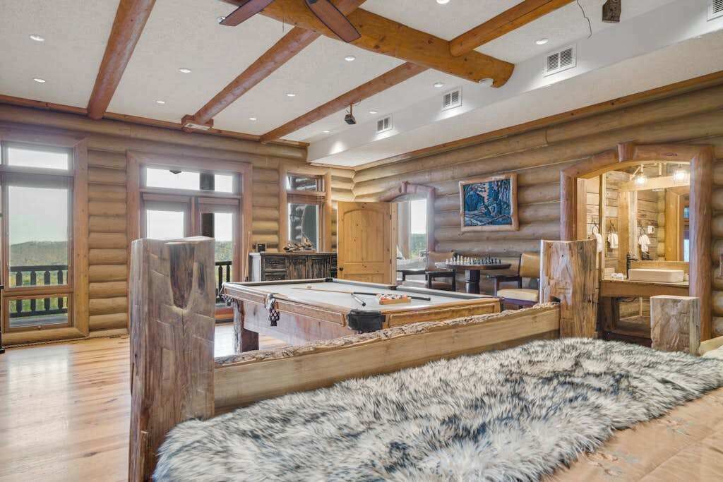 A log cabin interior with a pool table, wooden beams, large windows, and a fur rug.
