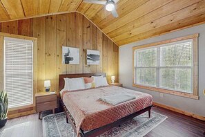 Un dormitorio con techo de madera, cama con cubrecama acolchado, dos marcos de cuadros en la pared, una mesita de noche con lámpara y un ventanal con persianas.