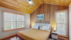 A bedroom with a wooden ceiling, a bed with a headboard, two bedside tables, and a painting on the wall. - Dwell and Wander Stay (Ellijay)