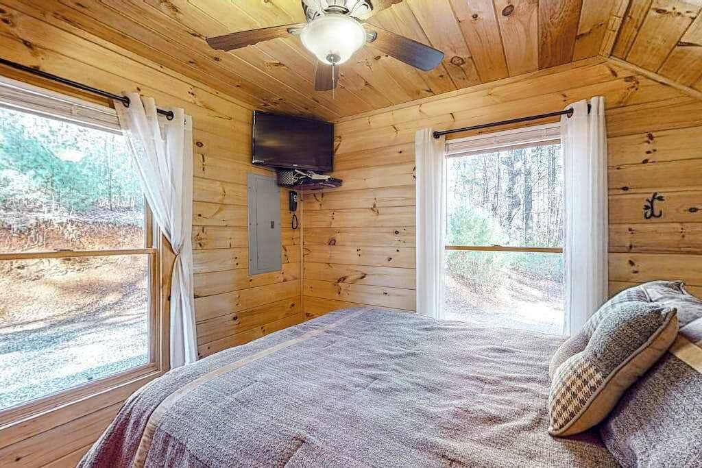Camera da letto in una baita di legno, con un letto, una ventola a soffitto, una TV a parete e ampie finestre.