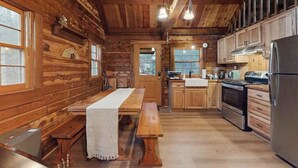 Interior de una cabaña de madera con una mesa de comedor, bancos, una zona de cocina con electrodomésticos y ventanas amplias.