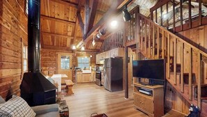 Interior de una cabaña de troncos con chimenea, escalera de madera, cocina y televisor.