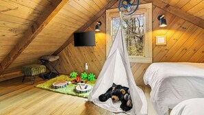 Ein gemütliches Schlafzimmer im Dachgeschoss mit Holzbalkendecke, einem Bett, einem Hängesessel, einem Fernseher und einem Fenster mit Blick auf Bäume.