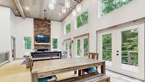 A spacious dining area with a large wooden table, a stone accent wall, and a TV mounted above the fireplace. - The Hidden Haven Stay (Blue Ridge)
