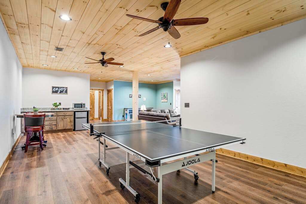A room with a ping pong table, ceiling fans, a kitchen area, and a seating area with a couch.
