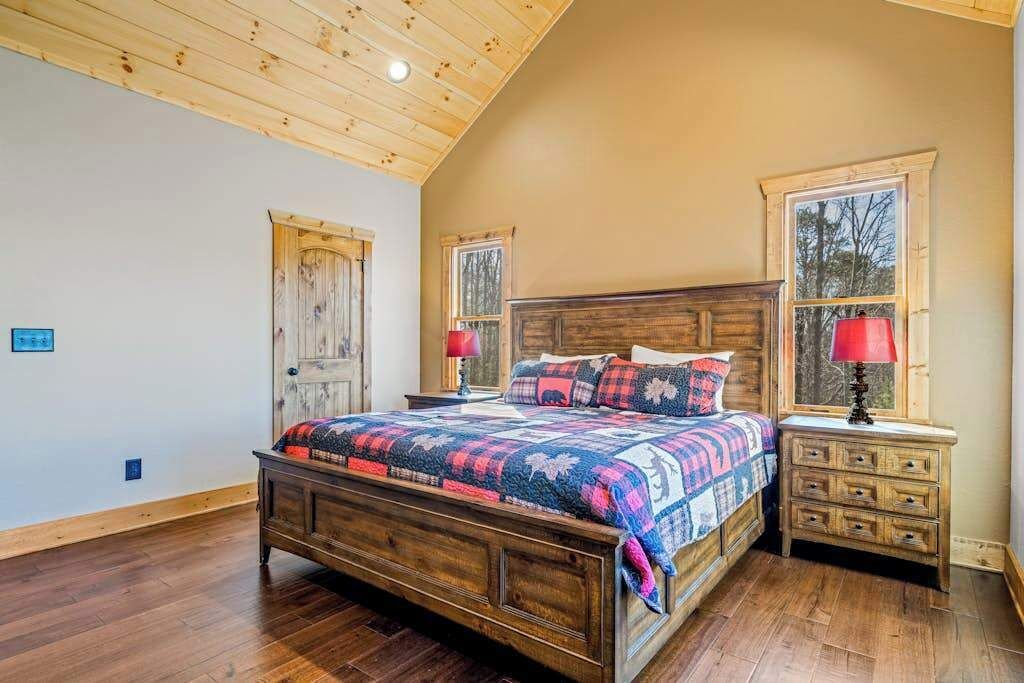 A bedroom with a wooden ceiling, a large bed with a colorful quilt, two bedside tables with lamps, and a window with a view of trees.
