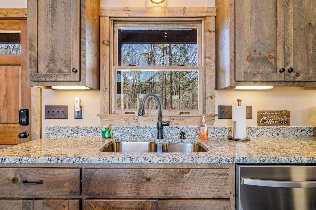 A kitchen with wooden cabinets, a granite countertop, a stainless steel sink, and a window with a view of trees.