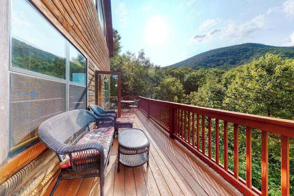 Una terraza de madera con mobiliario de mimbre, un cerramiento de vidrio y vistas a una montaña boscosa.