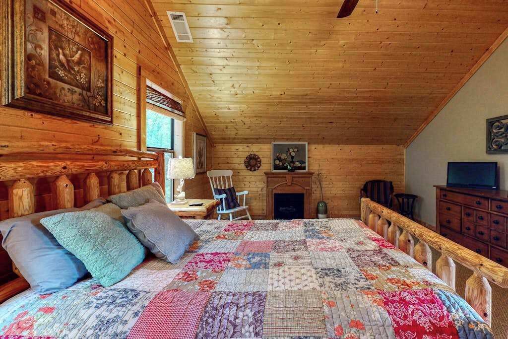 Et hyggeligt sovev ærelse i en hytte med træloft, en stor seng med blomsterquilt, en pejs og et fjernsyn.