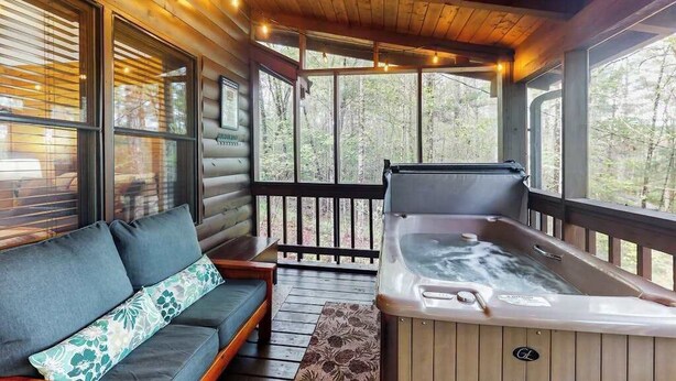 A log cabin with a wooden deck, a hot tub, and a sofa with a floral cushion. - Bluebird Bliss Cabin (Blue Ridge)