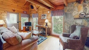 A log cabin interior with a sofa, armchair, stone wall, television, and a fireplace.