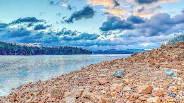 A rocky shoreline with a calm body of water, distant trees, and a cloudy sky.