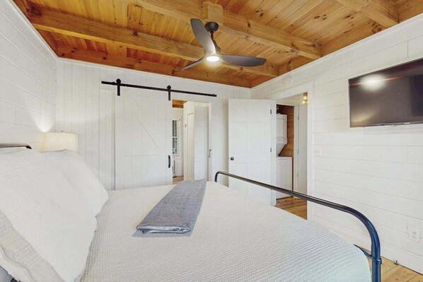 Un dormitorio con cama, ventilador de techo, televisor y puerta que da a otra habitación.
