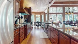 A kitchen with wooden cabinets, a stainless steel refrigerator, a dining area with a table and chairs, and large windows. - River Runs Through It (Dahlonega)