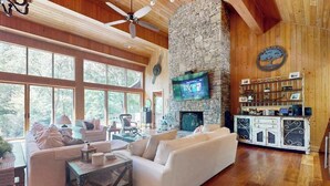 A spacious living room with a stone fireplace, large windows, and a television mounted on the wall. - River Runs Through It (Dahlonega)