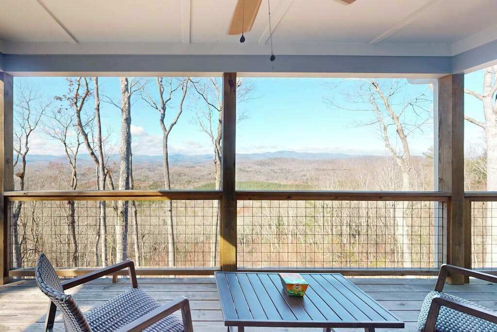 Uma área de estar coberta ao ar livre, com mesa e cadeiras, vista para árvores e colinas, e céu claro.