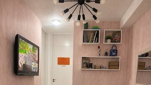 En stue med en brun sofa, et fjernsyn monteret på væggen, en loftlampe og en lille reol med pynteting.
