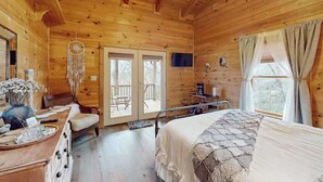 Quarto de cabana de madeira com cama, escrivaninha, cadeira, enfeite de pena, televisão e janela com cortinas.