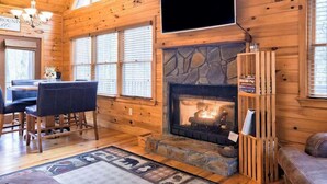 A wooden cabin interior with a stone fireplace, a dining area, and a television mounted on the wall. - Bear in the Woods Stay (Blue Ridge)