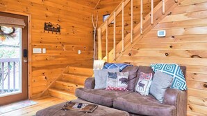 A wooden cabin interior with a sofa, a welcome sign, a fireplace, and a staircase. - Bear in the Woods Stay (Blue Ridge)