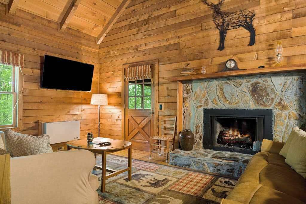Interior de uma cabana de madeira com lareira de pedra, paredes de madeira, televisão na parede e um espaço de estar acolhedor.