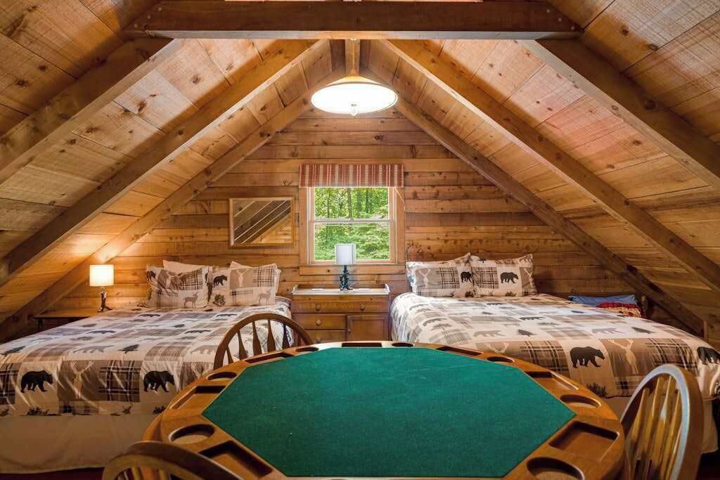 An attic bedroom with wooden beams, two beds with animal-themed bedding, a green table, and a window with a view of greenery.
