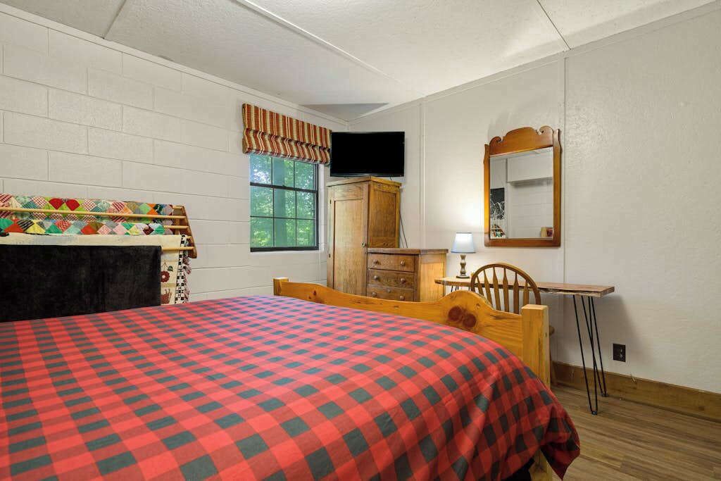 A bedroom with a bed, a wooden dresser, a mirror, a window with curtains, and a television.