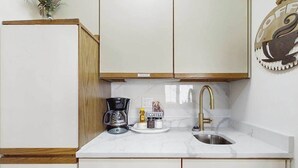 A kitchen with white cabinets, a coffee maker, a sink, and a coffee-themed wall decoration. - Cool River Condo I Stay (Helen)