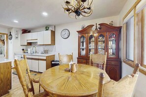 Een eetruimte met een ronde houten tafel, houten stoelen, een kroonluchter, een klok en een houten kast met glazen deuren.