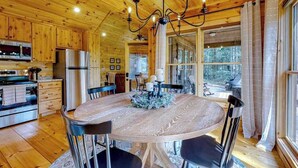 Interior de una cabaña de madera con una mesa de comedor, sillas, una lámpara de araña y una zona de cocina.