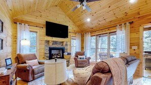 Interior acogedor de una cabaña con chimenea, sofás de cuero, televisor y ventanas grandes.