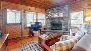 A wooden cabin interior with a stone fireplace, a television, and plaid furniture. - Ogana Ridge Getaways (Ellijay)