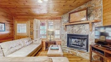 L’intérieur chaleureux d’un chalet avec une cheminée en pierre, des murs en bois, un canapé en cuir et un téléviseur.