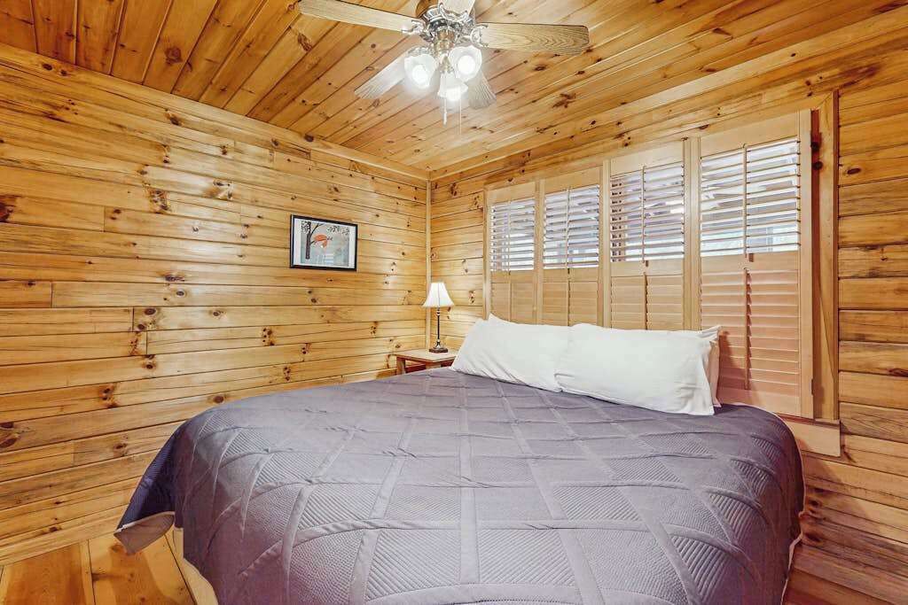 A wooden-paneled room with a bed, a ceiling fan, and a window with blinds.