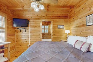 Une pièce de cabane en bois comprenant un lit, un téléviseur, un ventilateur de plafond et une fenêtre.