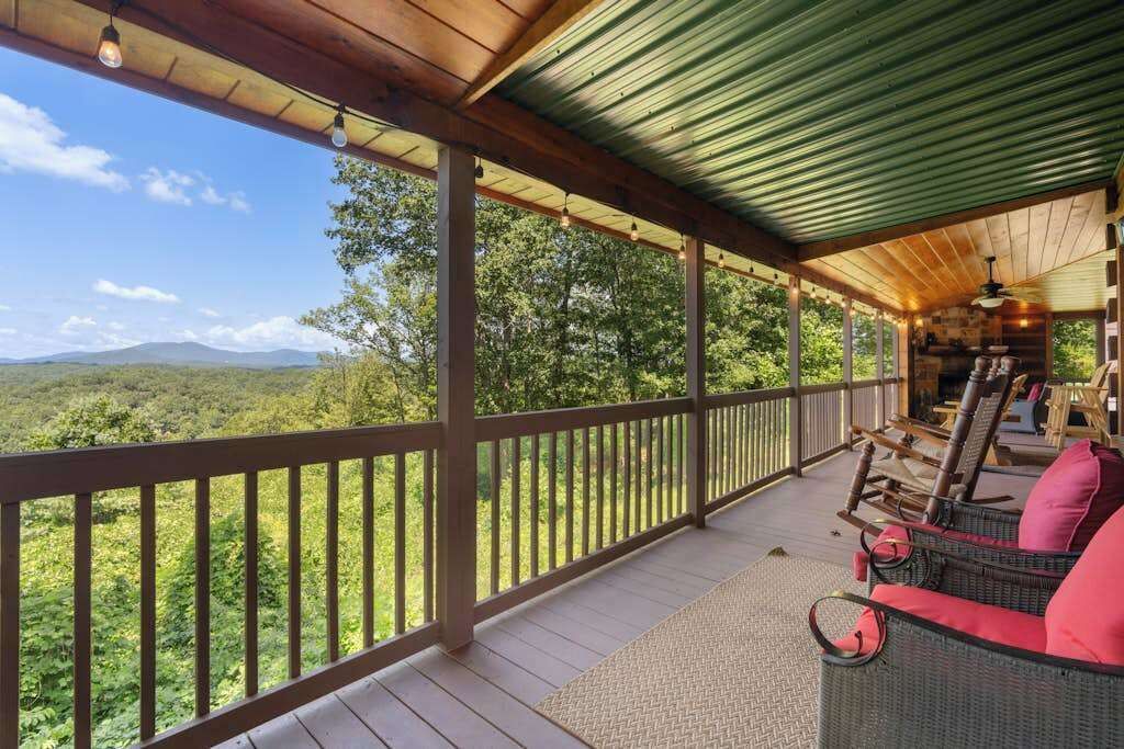 A covered porch with a view of a forest and mountains, wicker furniture, and a wooden ceiling.
