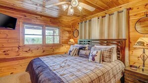 Camera da letto in una baita di legno, con un letto, una ventola a soffitto, una televisione e una finestra.