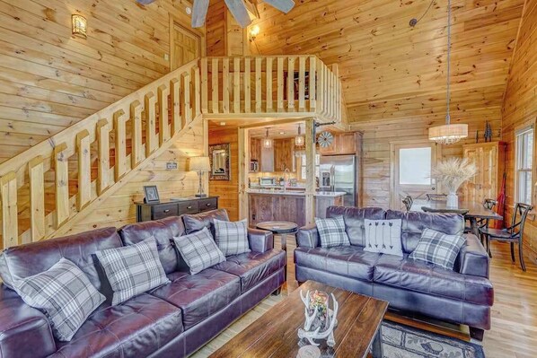 A wooden cabin interior with a staircase, two leather sofas with plaid cushions, a dining area, and a kitchen. - Cardinal House Retreat (Mineral Bluff)