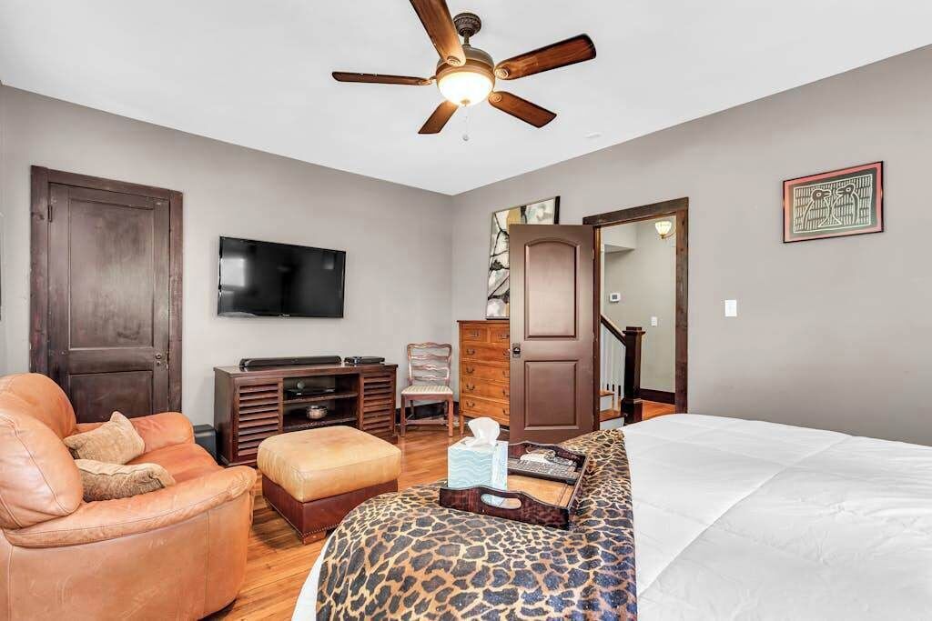 A bedroom with a bed, a television, a ceiling fan, a door, a chair, and a dresser.